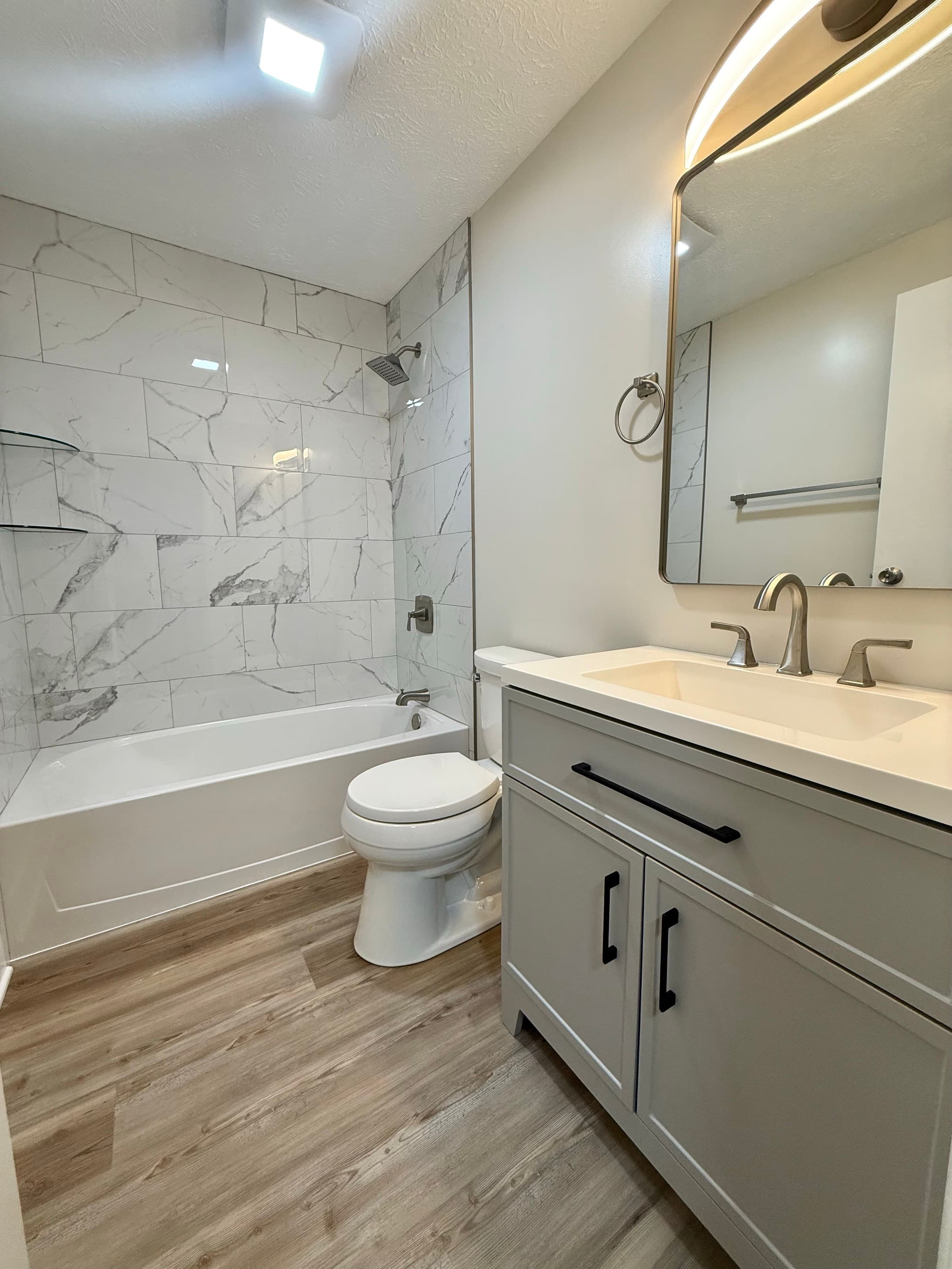 Remodeled bathroom with a marble-tiled tub surround, gray shaker vanity, quartz top, and brass-framed mirror