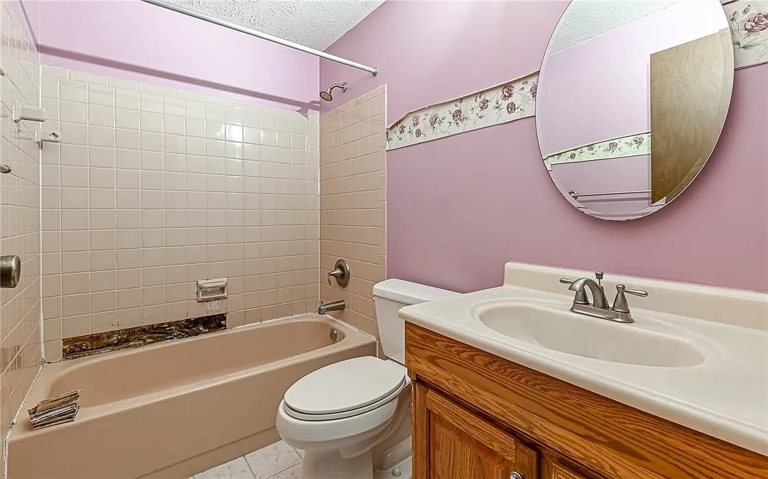 Dated bathroom with a pink floral wallpaper border, oak vanity, and worn tile before remodeling