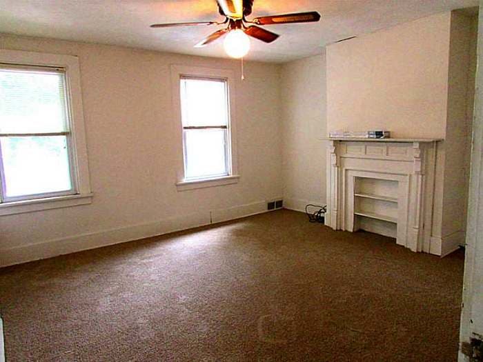 Living room with a ceiling fan, white mantel, and dark carpet before renovation