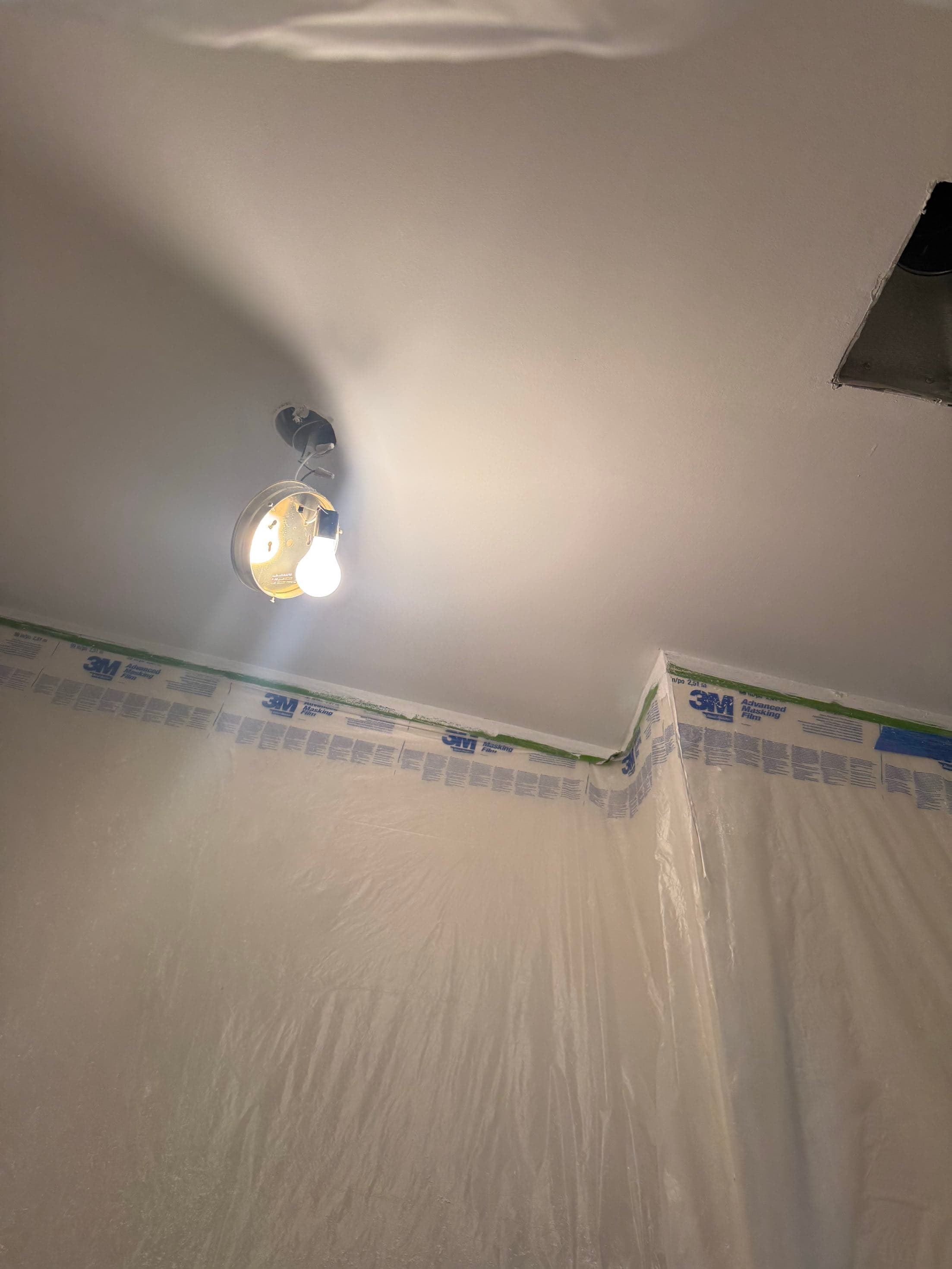 Close-up of a smoothed, freshly painted ceiling corner and sconce with masking film protecting the walls below