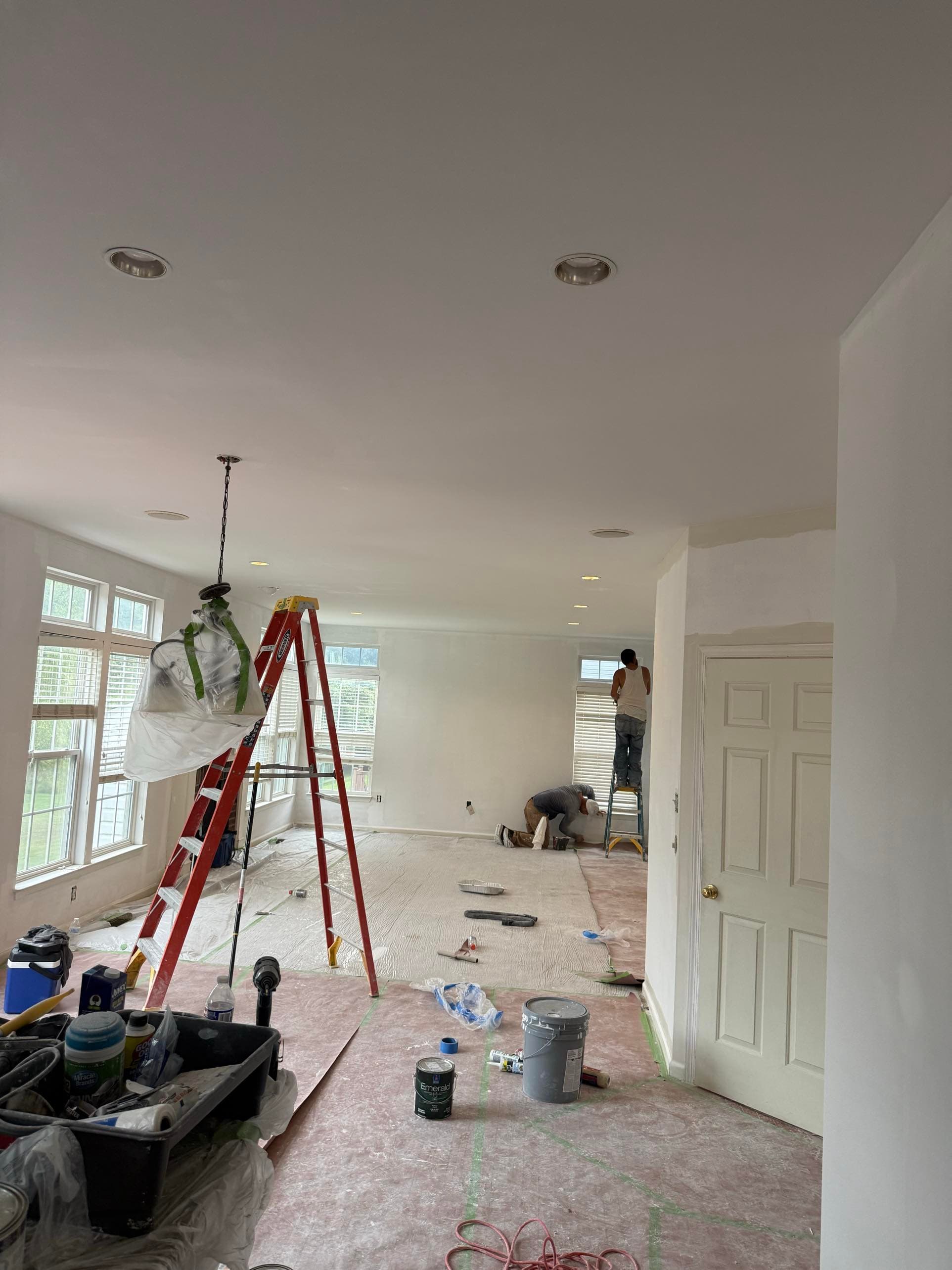 Two crew members prepping and painting the ceiling in an open living space with floors fully covered